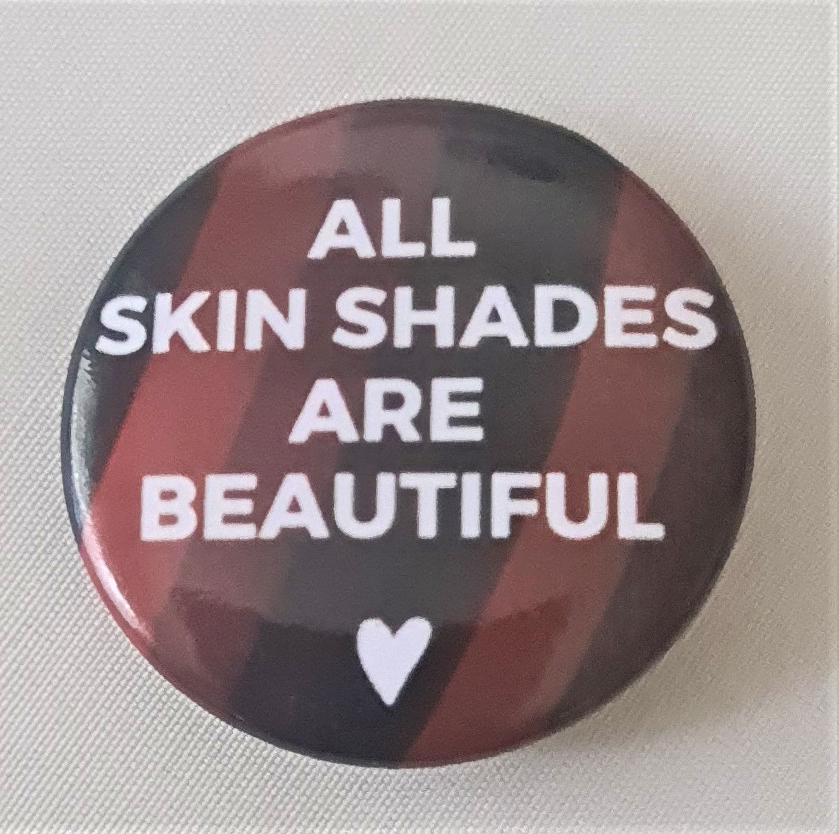 Round badge with embrace shades colours in the background, with the words, All skin shades are beautiful. and a white love heart below. 